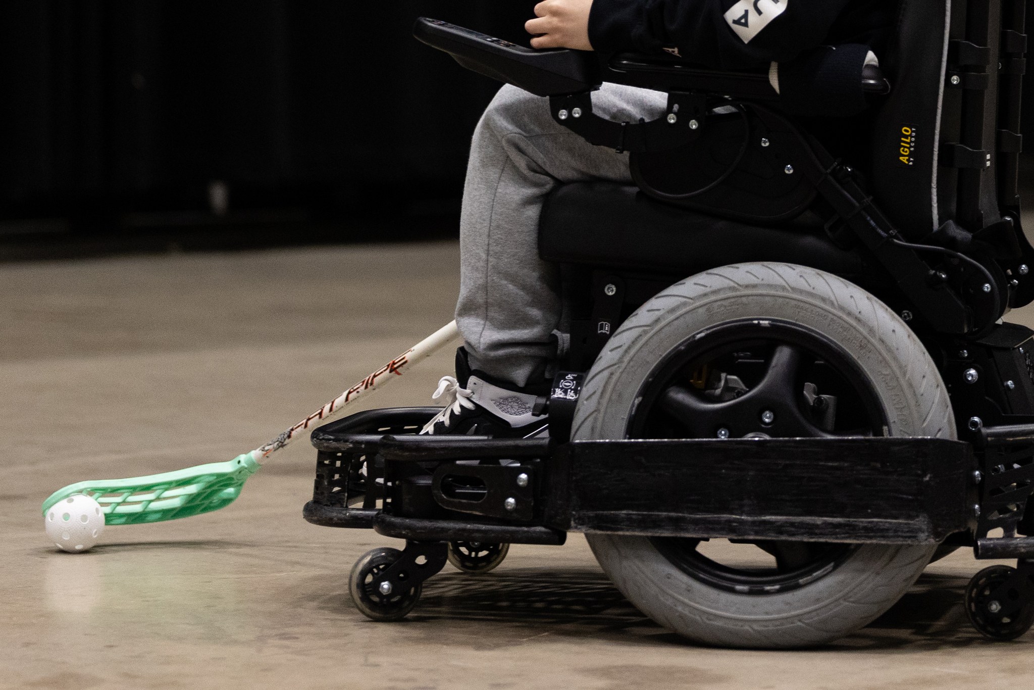 Lähikuvassa sisäpelikentällä kulkeva pelisähköpyörätuoli, jota käyttävä henkilö kuljettaa salibandymailalla palloa. 