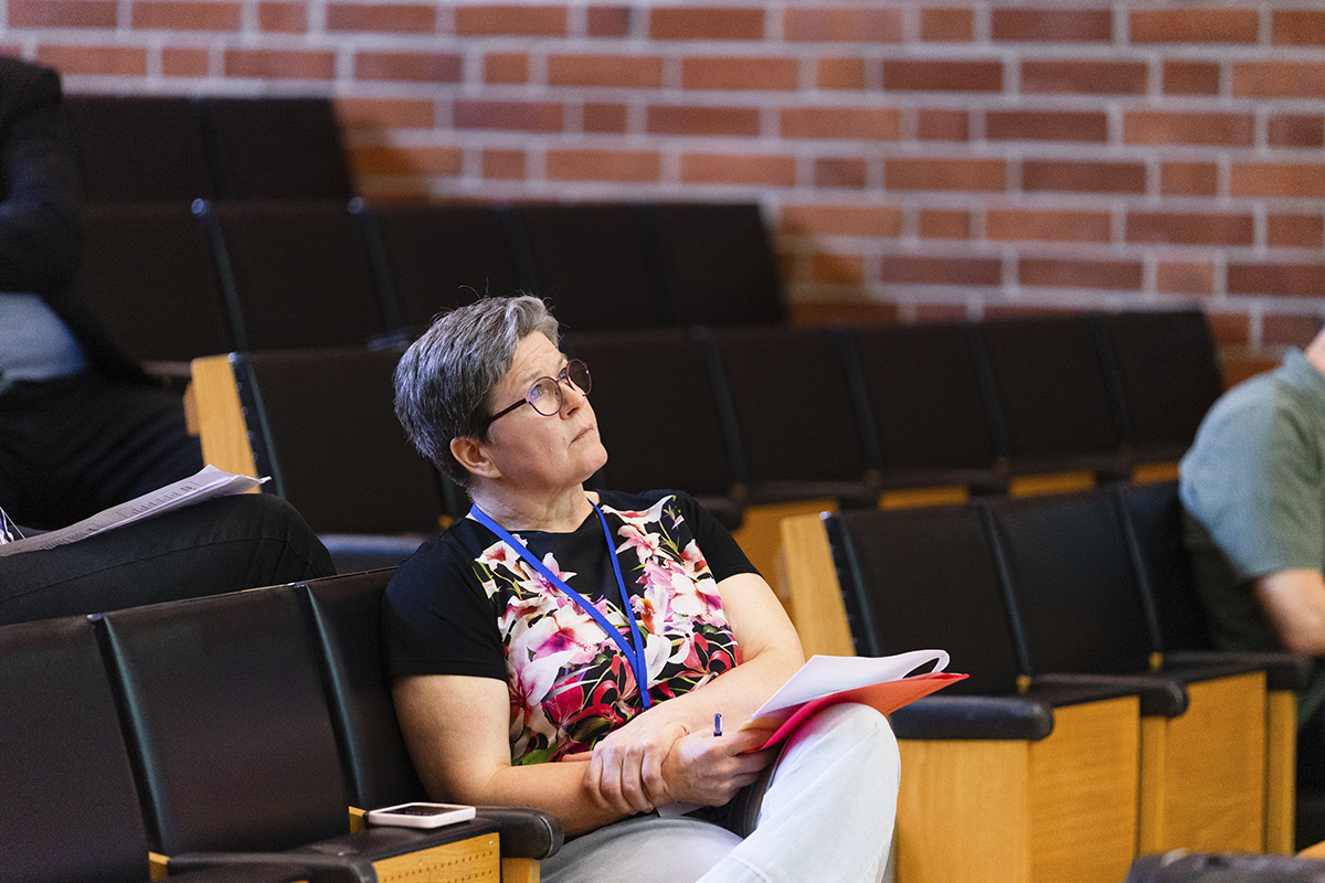Aija istuu muistiovihko sylissään tiiliseinäisen auditorion nousevan katsomon etuosassa, keskittyneen pohtivasti yläviistoon katsoen.