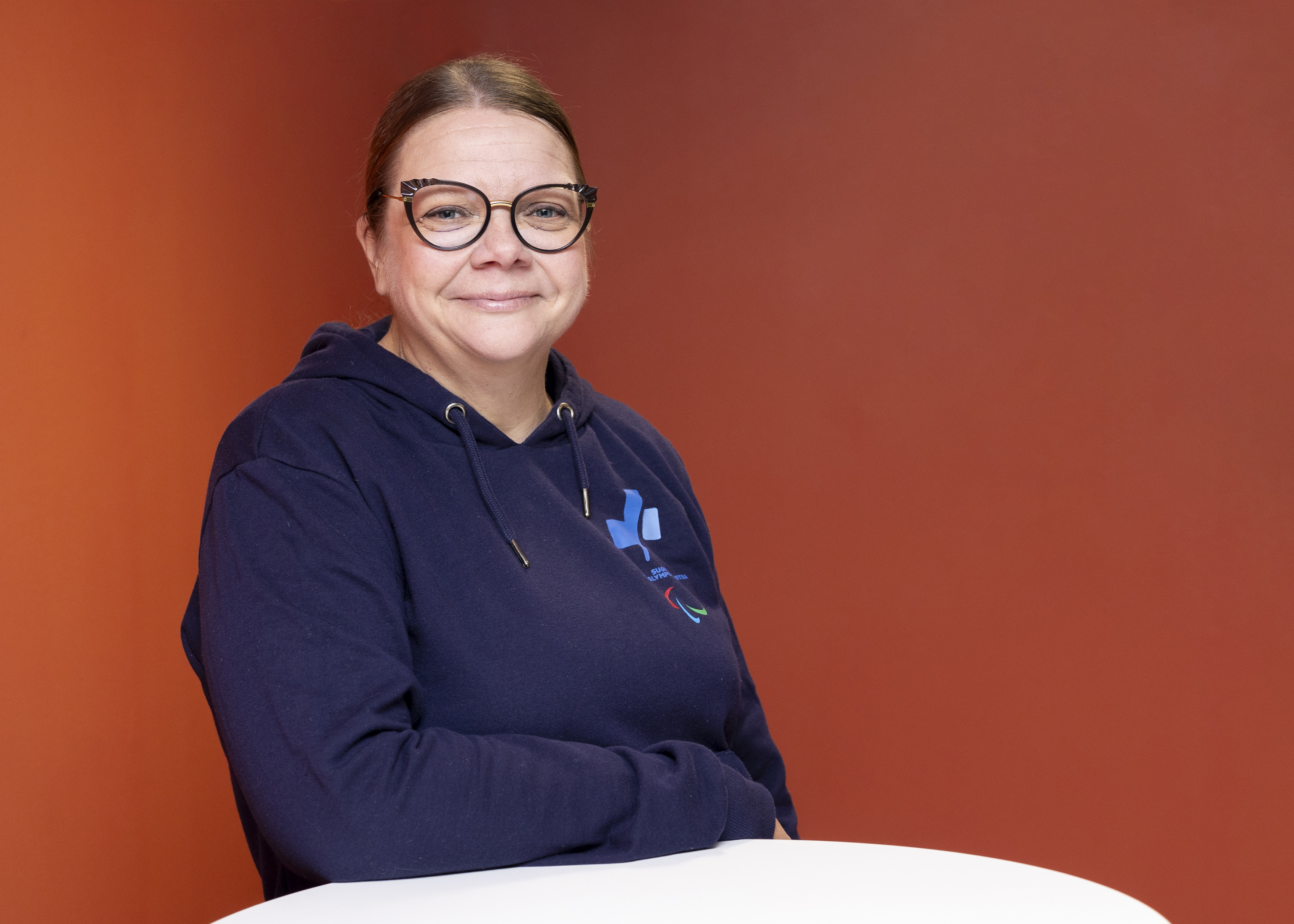 Katja Saarinen hymyilee Paralympiakomitean hupparissa kyynärvarsi edessä olevaan pöytään nojaten, punaisen taustan edessä.