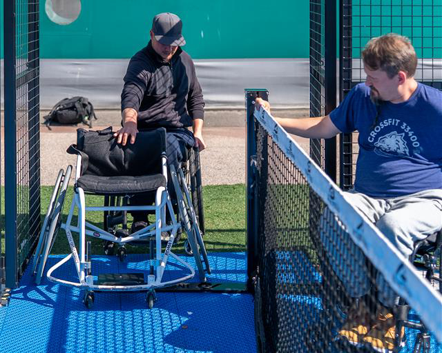 Pyörätuolia käyttävä henkilö tulossa sisään esteettömälle padelkentälle, erillistä pelituolia edessään sisään työntäen. Toinen pyörätuolia käyttävä pelaaja odottaa keskiverkon toisella puolella.