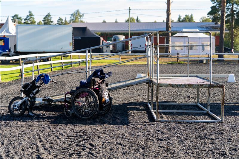 Liuskallinen teline ja nojapyörä aidatulla hiekkakentällä ulkona, taustalla parkkipaikalla telttoja ja hevostrailereita.