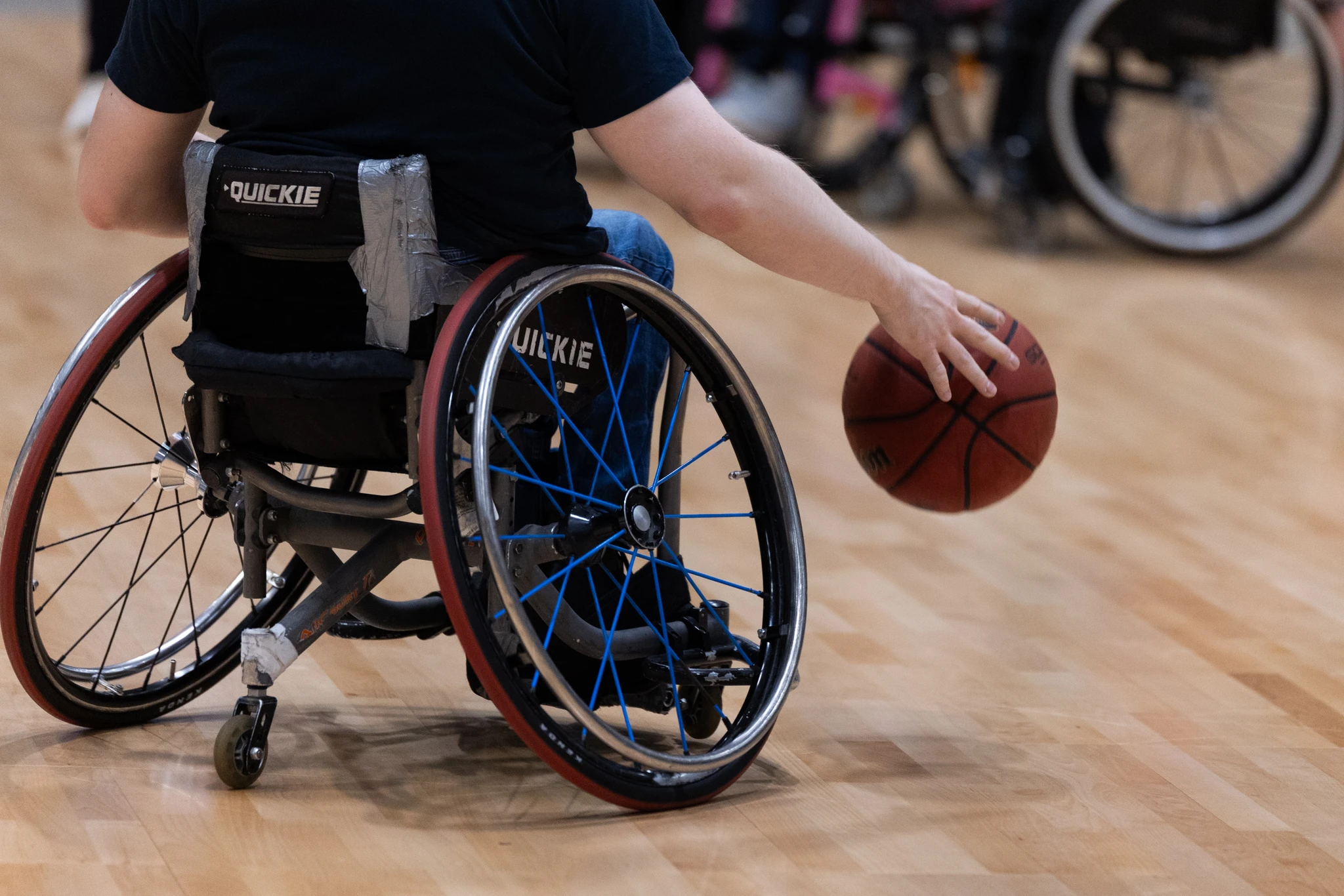Lähikuvaa pyörätuolikoripalloilijasta liikkumassa pallon kanssa. Kuva takaapäin, lapaluista alaspäin.