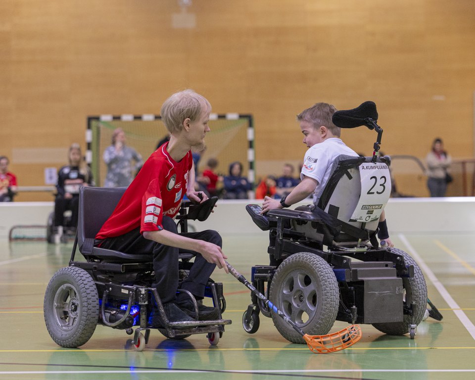 Kaksi spt-salibandypelaajaa (miesoletetut) kamppailemassa pallosta. Toinen punapaitainen ja toinen valkopaitainen.