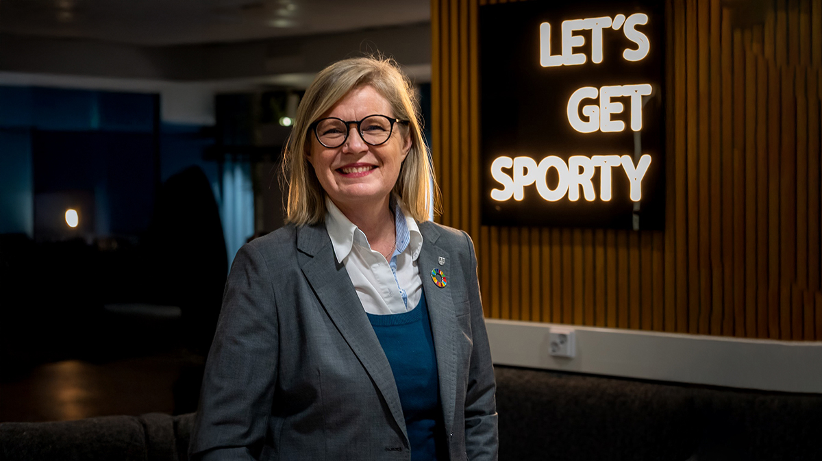 Sari Rautio hymyilee kisahallissa hämärässä iltaympäristössä. Taustalla valotaulu, jossa lukee "Let's get sporty".