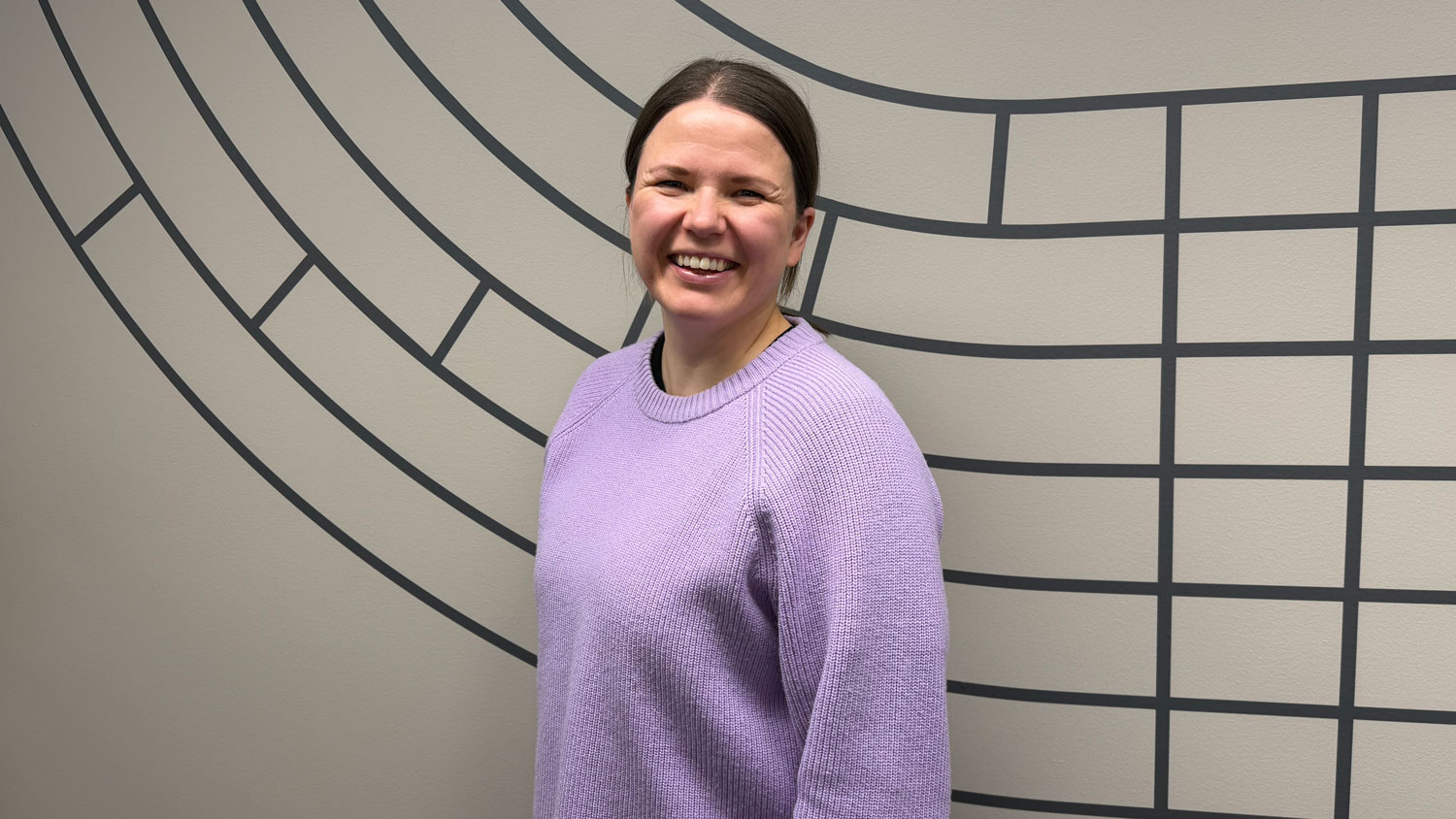 Heli Valtiala hymyilemässä kameralle vaaleanvioletissa villapaidassa, ruskeat hiukset kiinni. Taustalla graafinen seinäkuvio.