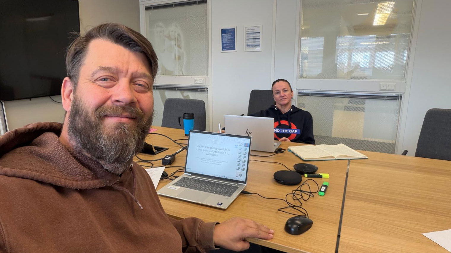 Jukka Parviainen ja Tiina Siivonen iloisessa selfiessä ennen webinaarin alkua. Kokoustila, läppärit ja muita työvälineitä pöydällä. 