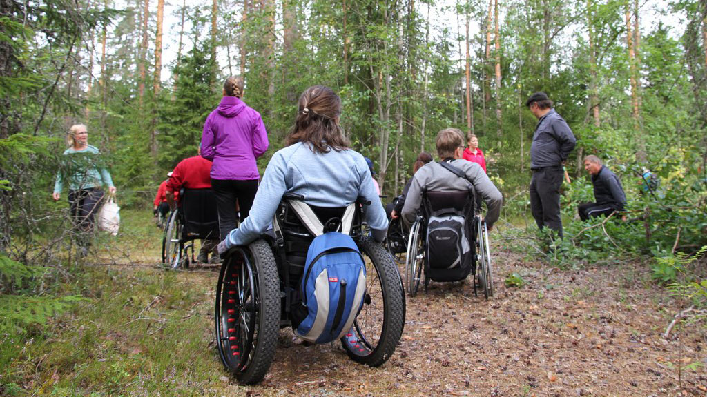 Henkilöitä maastopyörätuoleissa metsässä retkeilemässä.